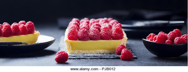 Raspberry tart with vanilla custard and white chocolate on slate board. Black stone background. Copy space. - Stock Image