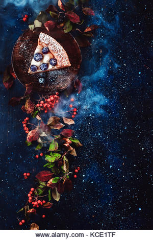 Floral border with a piece of blueberry pie, autumn leaves, berries and blue smoke on a dark wooden background. - Stock Image