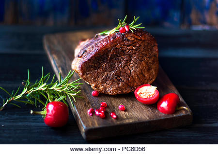 baked meat with rosemary and red pepper. steak. beef. dinner for men. dark photo. Black background. wooden board - Stock Image