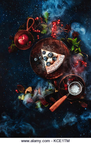 A piece of cake with powdered sugar and a strainer on a dark wooden background with floral decoration. Handmade - Stock Image