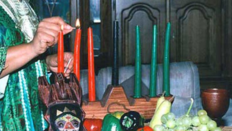 A woman lights three red candles, three green candles, and one black candle that are placed on a kinara