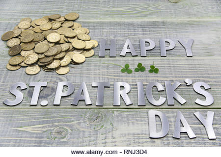 inscription happy Patricks day, a lot of gold British coins and three sheets of clover, lie on a textured wooden Board - Stock Image