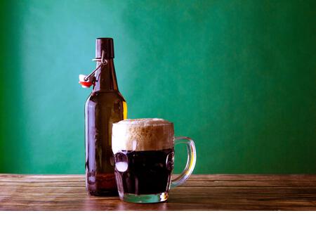 A mug of dark beer with its open bottle on a green background and a wooden table. St. Patrick and stout concept - Stock Image