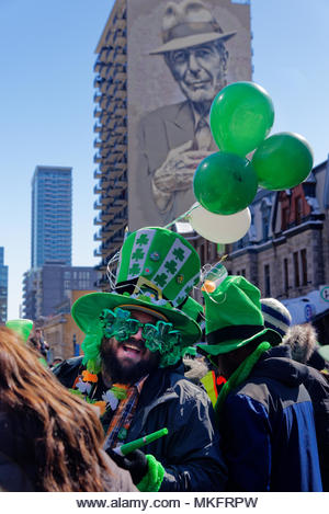 A laughing man wearing shamrock shaped sunglasses at the Montreal St Patricks Day parade, with the big portrait of Leonard Cohen behind - Stock Image