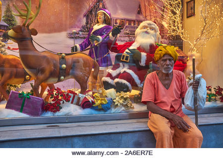 A turbanned elderly man, apparently from the Indian countryside, rests at a shop window displaying Christmas decoration; Colaba, Mumbai, India - Stock Image