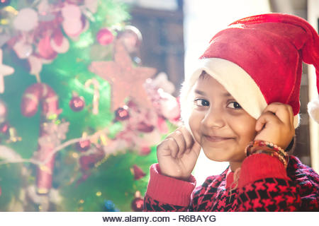 Little Indian/Asian kid girl in santa hat with Christmas tree and lights on background. - Stock Image