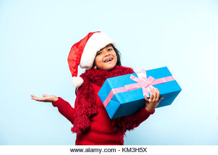 Little Indian girl holding christmas gifts in red cloths or santa clause hat and attire, standing isolated over - Stock Image