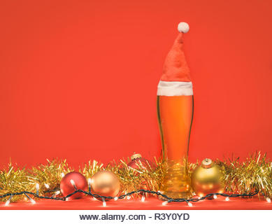 Close-up shot of full pilsner glass of pale lager beer or ale with Santa Claus or christmas red hat, christmas lights baubles and tinsel on red backgr - Stock Image