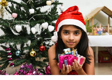 cute little Indian girl celebrating Christmas in India many present Indian Christmas tree. Happy child wearing Santa helper red hat with lot of x-mas - Stock Image