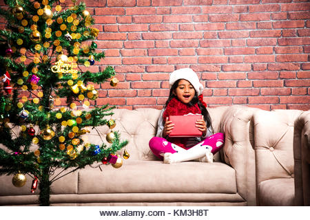 Little Indian girl holding christmas gifts in red cloths or santa clause hat and attire, standing isolated over - Stock Image