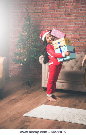 Little Indian girl holding christmas gifts in red cloths or santa clause hat and attire, standing isolated over - Stock Image