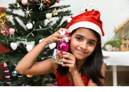 cute little Indian girl celebrating Christmas in India many present Indian Christmas tree. Happy child wearing Santa helper red hat with lot of x-mas - Stock Image