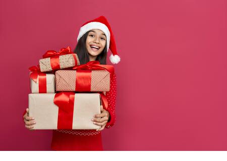 Excited indian kid girl wear santa hat hold many gifts boxes on red background. - Stock Image