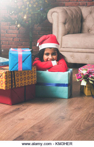 Little Indian girl holding christmas gifts in red cloths or santa clause hat and attire, standing isolated over - Stock Image