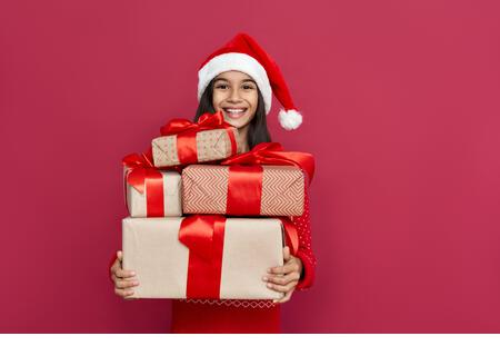 Happy indian kid girl wear santa hat holding many gifts boxes on red background. - Stock Image