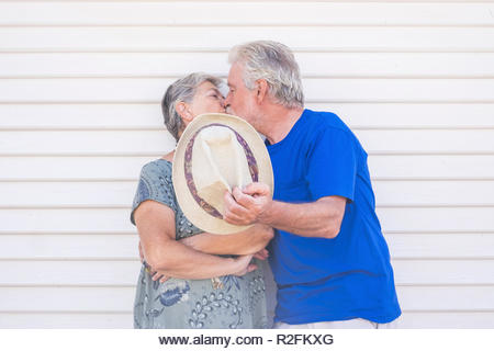 Valentine's day concept with elderly beautiful senior couple kissing hidding with a beige hat with a white wood background wall behind them. love and forever life together with caucasian couple man and woman happy - Stock Image