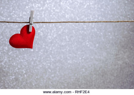 Single red heart hanging from string by clothes peg on silver background. Romantic Valentine's Day scene with copy space - Stock Image