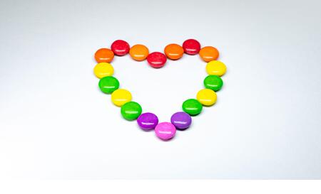 Selective focus of colorful button chocolates arranged in the shape of a heart on a white paper background. - Stock Image