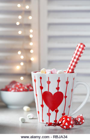 Hot chocolate with marshmallows, red heart on the cup, winter background with lights out of focus. Winter or Valentine's day background. - Stock Image