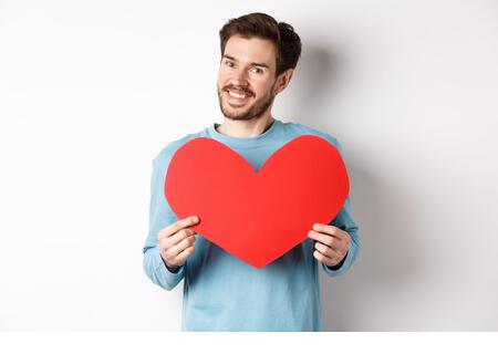 Handsome european man in sweater saying I love you, boyfriend standing with valentines day red heart, posing over white background - Stock Image