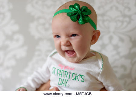 Irish baby with big blue eyes laughing and wearing St Patricks Day outfit and shamrock headband - Stock Image