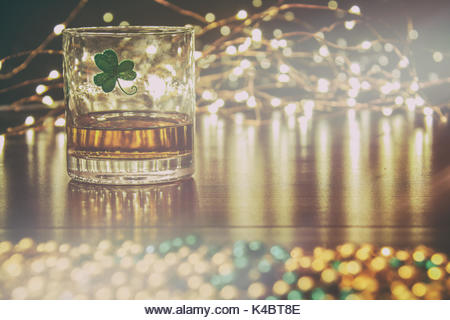 Irish whiskey in a glass with a clover symbol, on a pub table with gold beads and bar lights. - Stock Image